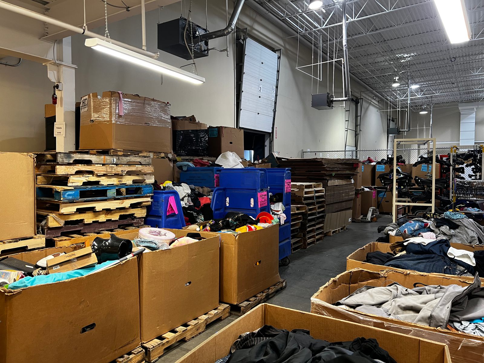 A Goodwill back room: pallets stacked with sealed boxes, open cardboard bins and blue bins overflowing with donated clothing and household items, and a loading door, empty hanger racks and flattened cardboard behind them.
