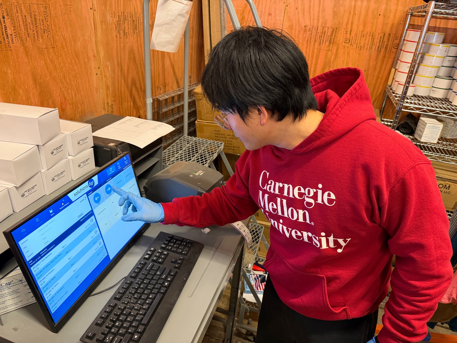 A person in a red Carnegie Mellon sweatshirt and blue gloves operates the Solutions POS touchscreen at a store workstation, with a label printer beside it.