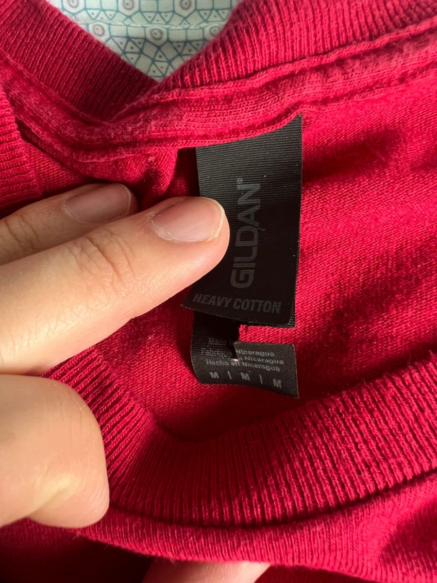 Fingers holding open the black care label at the collar: GILDAN, HEAVY COTTON, size M